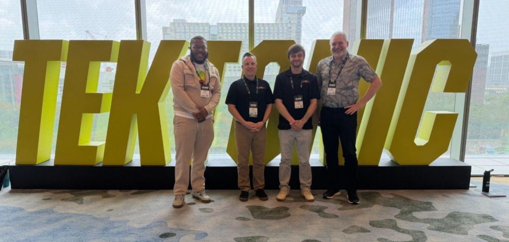 Four men with lanyards pose in front of oversized yellow letters indoors; city skyline visible through glass walls.