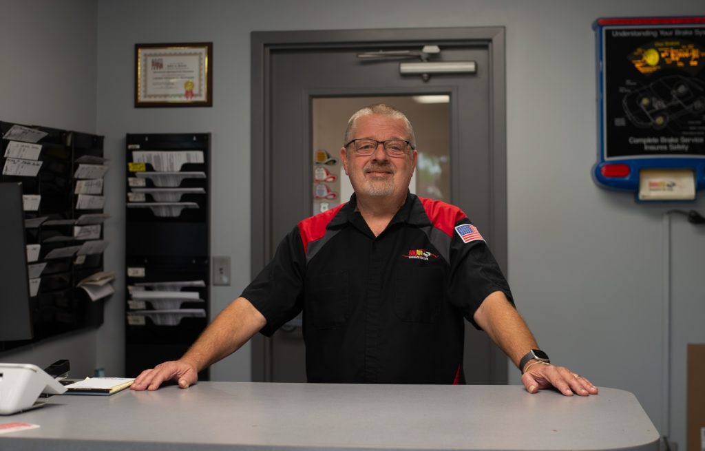 Male technician in a black and red uniform stands behind a gray reception counter in a workshop, smiling at the camera with shelves and tools in the background.