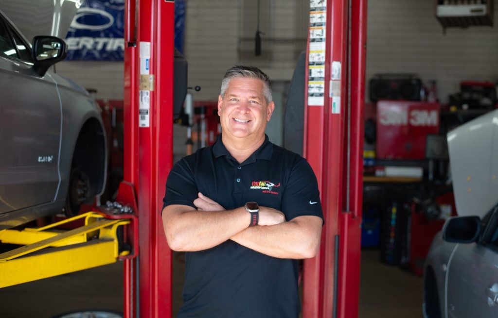 Smiling auto mechanic with arms crossed stands in a repair shop between red car lifts and cars nearby.
