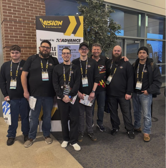 Eight men in matching black shirts with lanyards pose together at a trade show in front of a Vision banner with yellow chevrons.