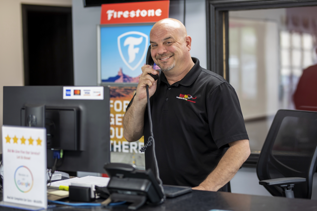 Man at a service desk inside an auto shop talks on a coiled telephone cord, with a Firestone sign and window in the background.