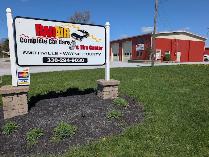 Exterior view of a RAD AIR Complete Car Care & Tire Center sign in a landscaped bed, with a red industrial building in the background and a blue sky.